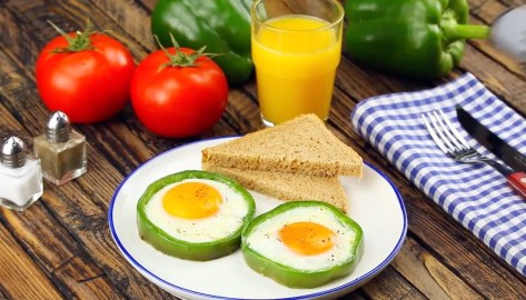 Ideen fürs Frühstücksei: 2 Eier im Paprikaring neben Toastscheiben auf einem Teller. Daneben steht ein Glas Orangensaft und im Hintergrund liegen frische Zutaten.