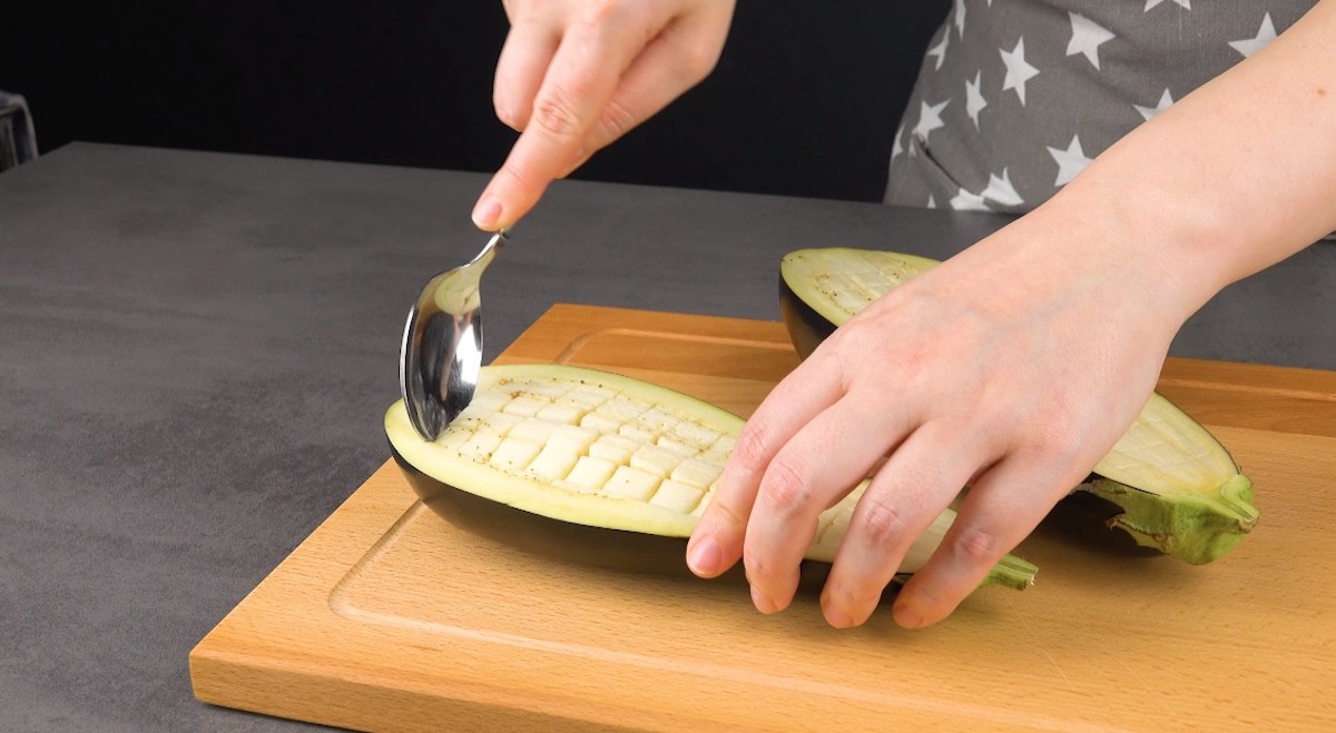 Eine halbierte Aubergine wird rasterförmig eingeschnitten und mit einem Löffel die Fruchtwürfel entfernt.