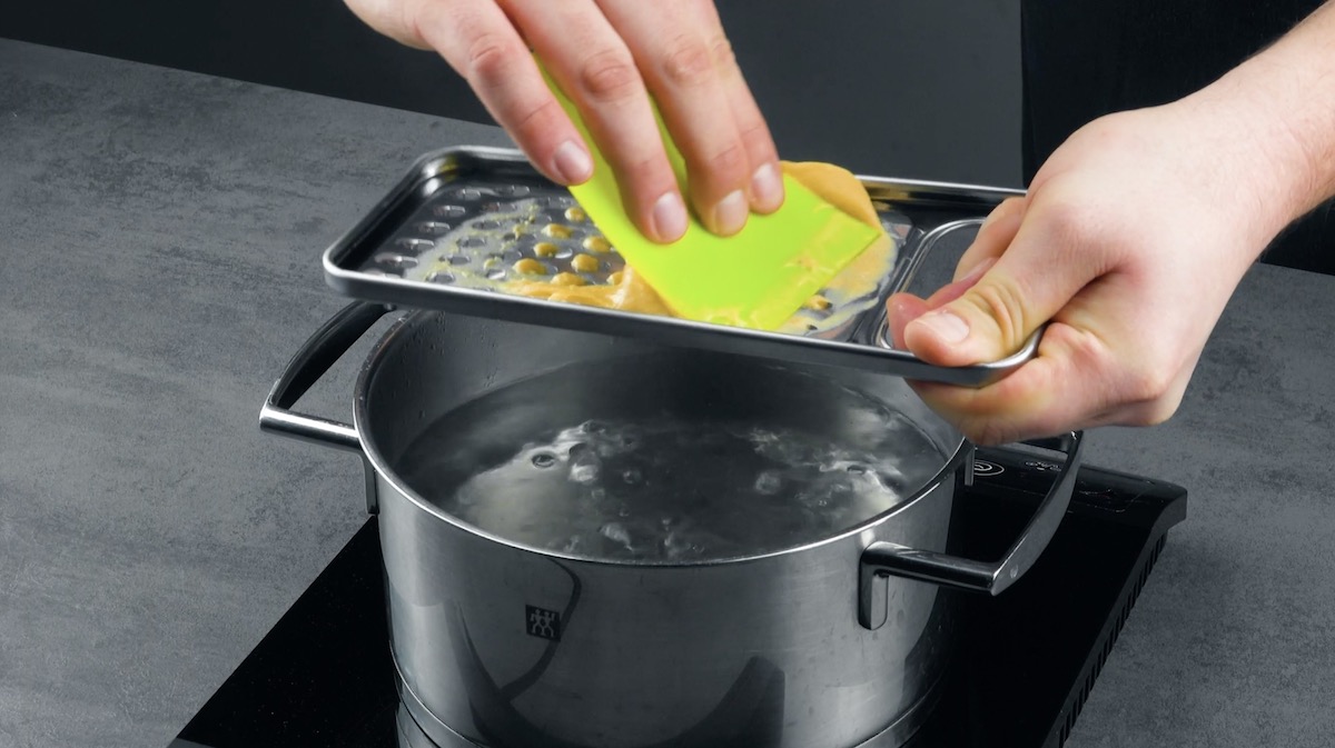 Spätzleteig wird mit einer Reibe und einem Teigschaber in einen Topf mit Wasser gerieben