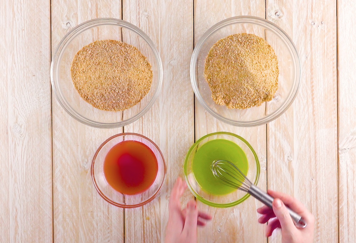 Zerbröselte Butterkekse in zwei Schüsseln neben einer Schüssel mit flüssiger Butter in Grün und einer Schüssel mit flüssiger Butter in Rot