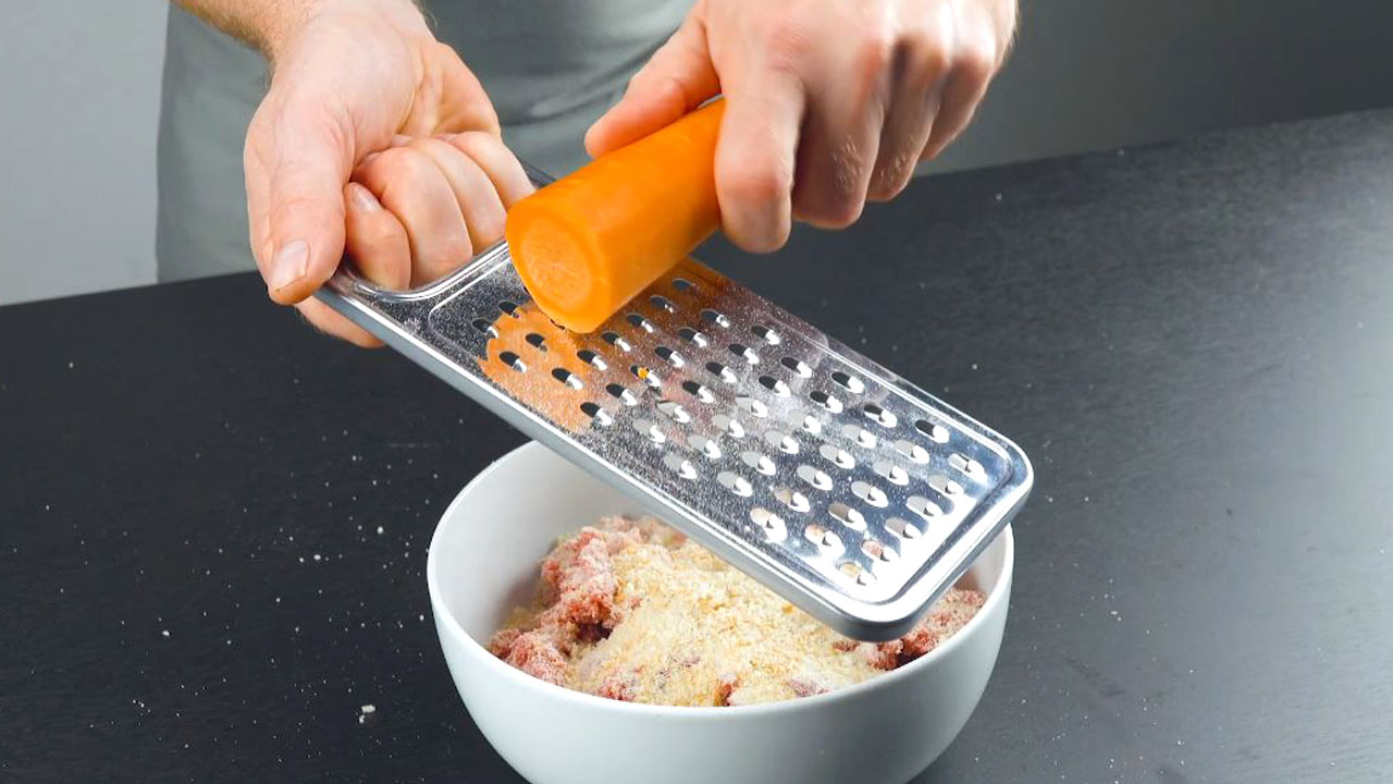 Karotte wird mit Reibe über Zutaten für Hackbällchen gerieben