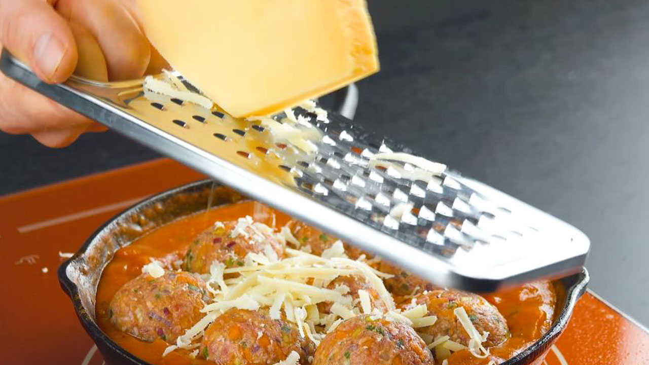 Käse wird mit Reibe über Hackbällchen in Pfanne gerieben