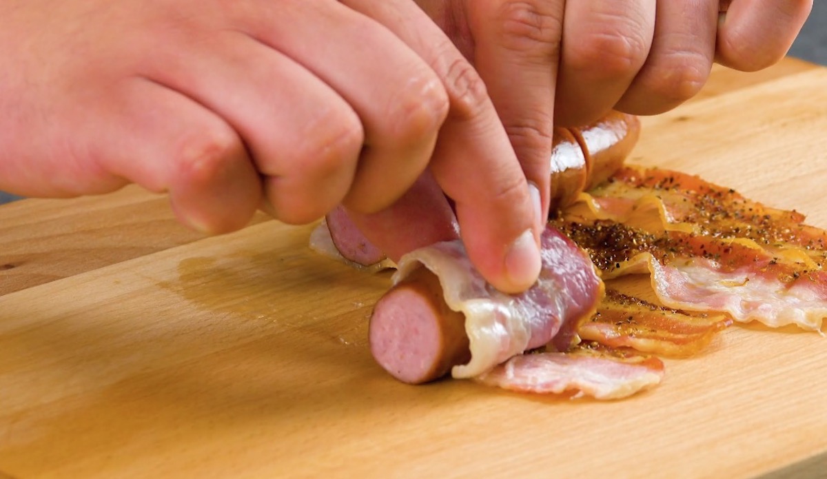 Halbiertes Würstchen wird in Baconscheibe eingewickelt