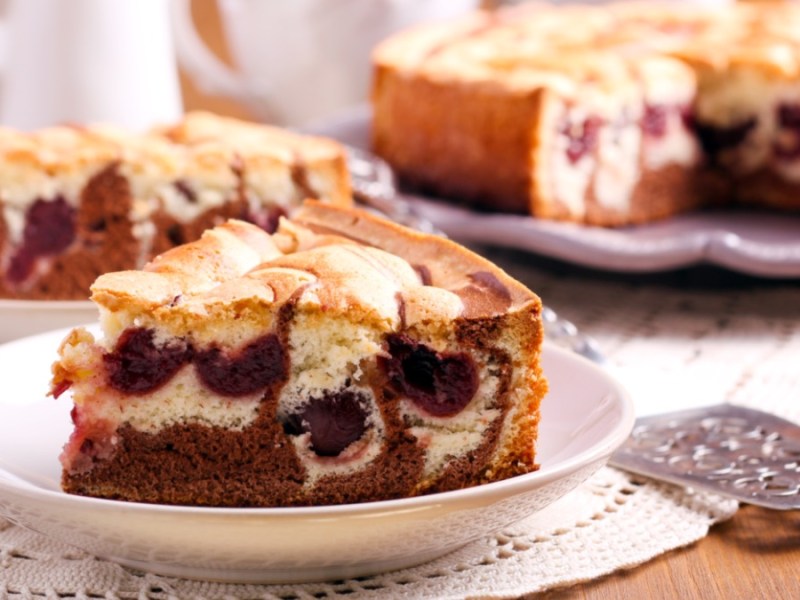Ein Stück gefüllter Marmorkuchen mit Kirschen auf einem Teller. Im Hintergrund sind der restliche Kuchen und ein weiteres Stück auf einem Teller zu sehen.