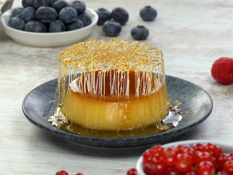 Ein Pudding mit Topping aus Karamell auf einem schwarzen Teller. Drumherum liegen frische Beeren.