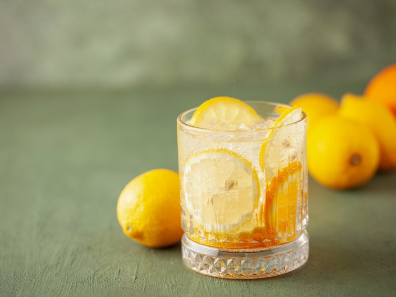Ein Glas Limoncello Tonic mit Eiswüfeln und Zitronenscheiben, dahinter frische Zitronen.
