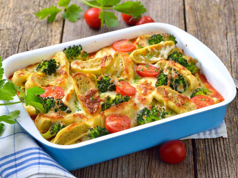 Maultaschen-Lasagne in einer blauen Auflaufform, dekoriert mit frischen Kräutern. Einige Kirschtomaten liegen neben der Schale.