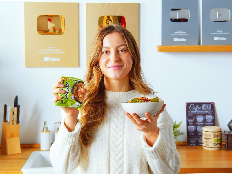 Eine Köchin hält eine Schale knuspriger Ramen-Salat und eine Packung Instant-Nudeln in die Kamera.