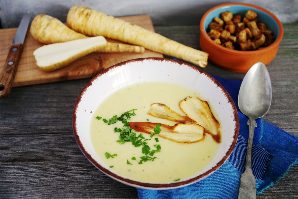 Pastinakensuppe in einer weißen Schüssel mit Pastinakenchips und Petersilie garniert. Daneben liegen Croutons und ganze Pastinaken.