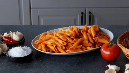 Penne Arrabiata, angerichtet auf einem Teller. Tomaten und Gewürze liegen außen herum.