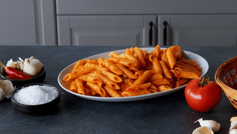 Penne Arrabiata, angerichtet auf einem Teller. Tomaten und Gewürze liegen außen herum.