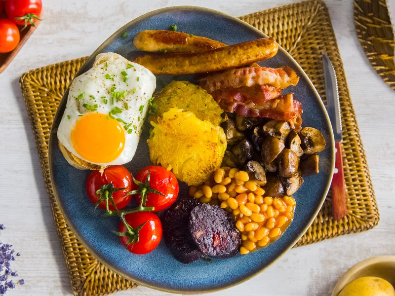 English Breakfast mit Bacon, Würsatchen, Spiegeleiern, Baked Beans, Tomaten, Pilzen und Toast auf einem weißen Teller. Daneben Messer und Gabel.