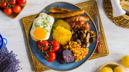 English Breakfast mit Bacon, Würsatchen, Spiegeleiern, Baked Beans, Tomaten, Pilzen und Toast auf einem weißen Teller. Daneben Messer und Gabel.