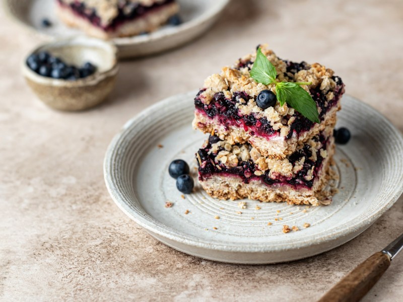 3 Frühstücksriegel mit Blaubeeren auf einen Teller gestapelt, dekoriert mit einem Minzblättchen, daneben eine Gabel., dahinter ein weiterer Teller.