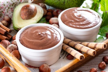 Zwei Schälchen mit Schokocreme aus Avocados. Drumherum liegen eine halbe Avocado mit Kern und Gebäckröllchen.