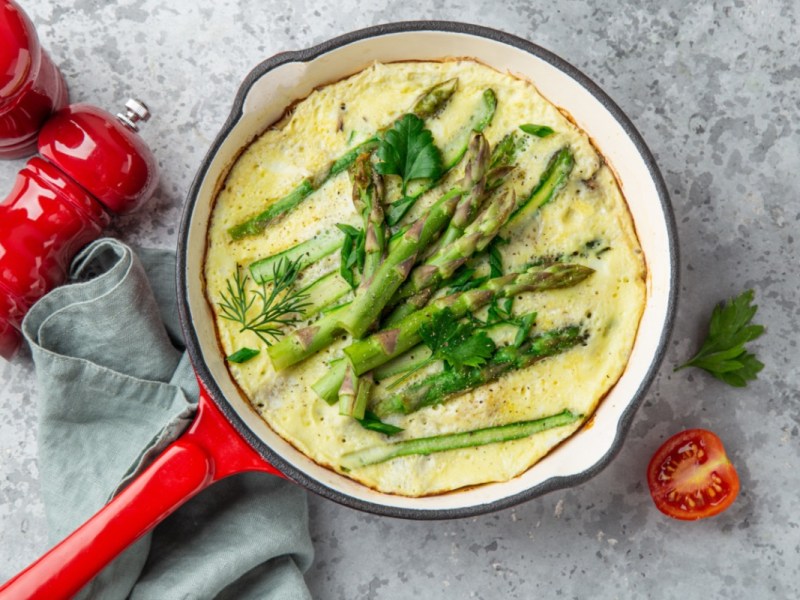 Ein Spargel-Omelett in einer roten Pfanne in der Draufsicht. Daneben liegen Pfeffer- und Salzmühlen in Rot.