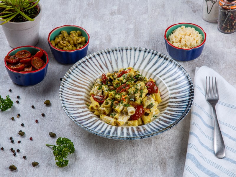 Italienischer Nudelsalat mit Tomaten und Kräutern auf einem Teller, daneben eine Gabel und Schüsseln mit Zutaten.