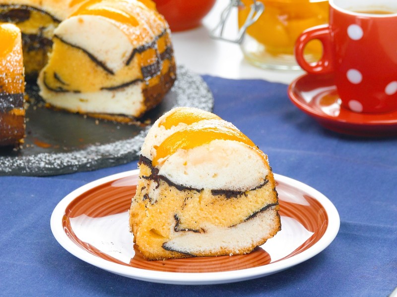 Ein Stück Pfirischkuchen mit Kakao im Wellenmuster auf einem Teller.