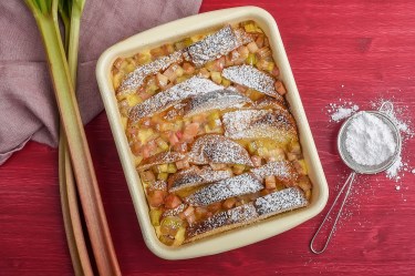 Auflaufform mit Brioche-Auflauf mit Rhabarber auf rotem Untergrund. Puderzucker und Rhabarber daneben.