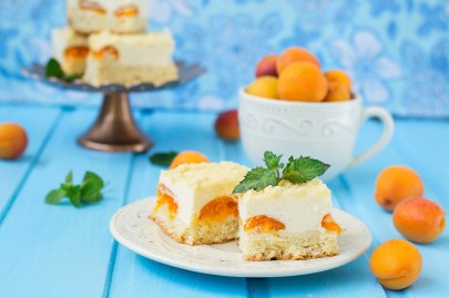 Einige Stücke Aprikosen-Schmandkuchen auf einem weißen Teller. Im Hintergrund unscharf weitere Stücke auf einem Kuchentablett sowie Aprikosen in einer großen Tasse.