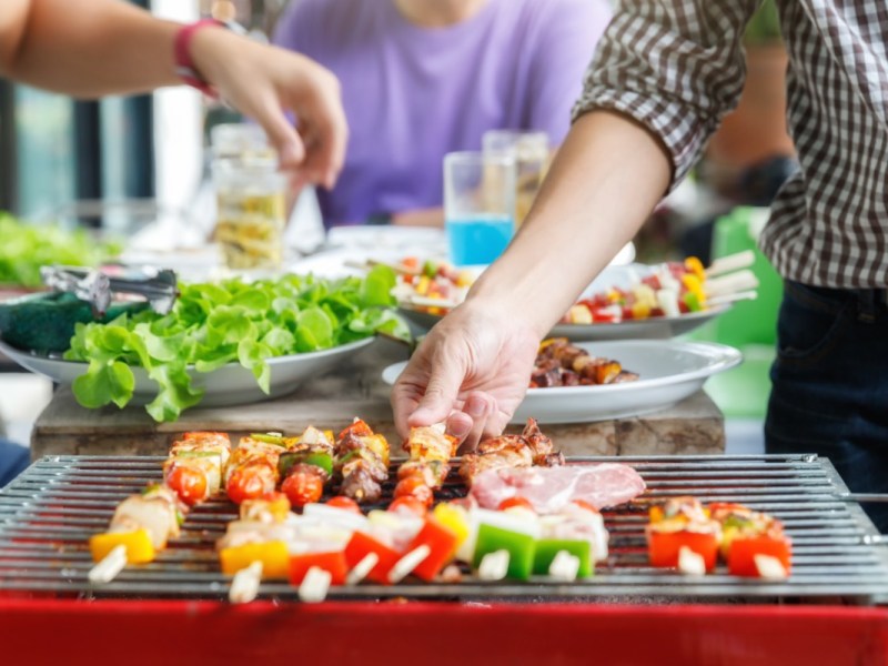 Was zu einem Grillabend dazu gehört: ein Grill mit Spießen im Vordergrund. Dahinter Beialgen und Menschen.