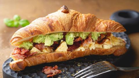 Ein Croissant mit Rührei Avocado und Speck auf einer Schieferplatte, davor eine Gabel.