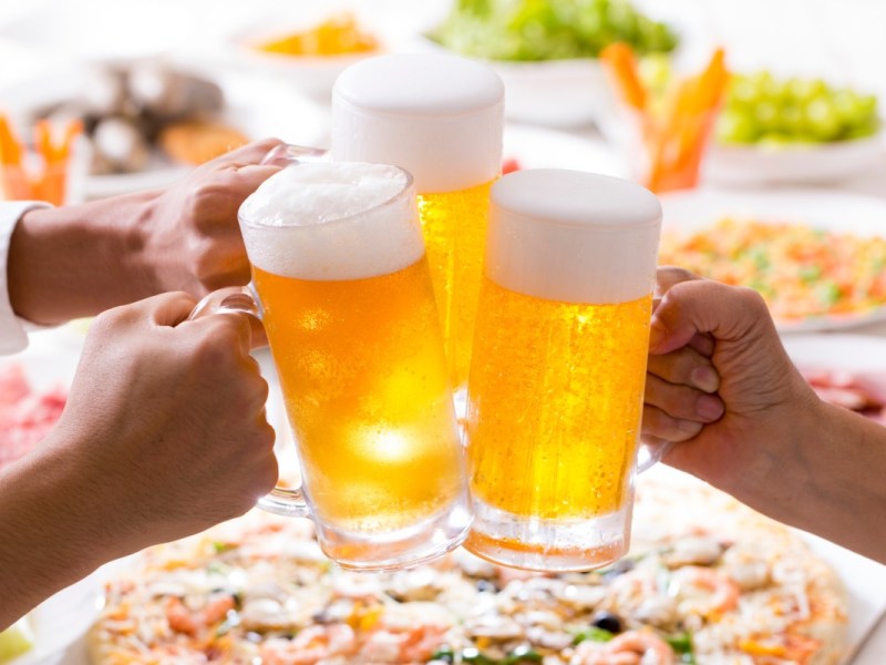 Tag des Biers: 3 Bierkrüge werden aneinander gestoßen. Im Hintergrund ist Essen zu sehen.