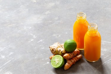 Zwei Flaschen Kurkuma-Shot. Daneben liegen Zutaten wie Ingwer, Kurkuma und Limetten.