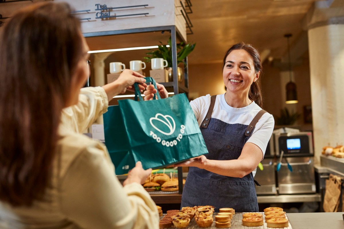 Eine dunkelhaarige Frau reicht einer anderen in einem Café eine Tüte mit der Aufschrit 