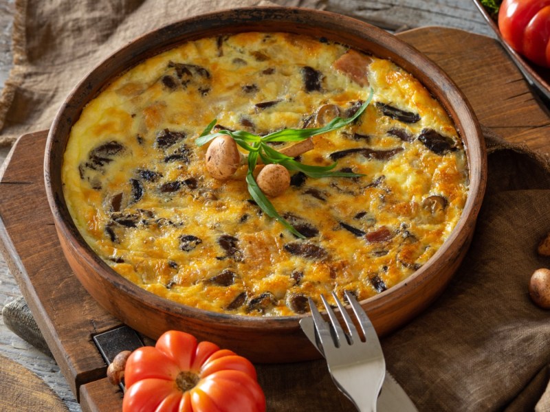 Eine Kartoffel-Frittata mit Pilzen in einer Pfanne. Drumherum liegen frische Zutaten.