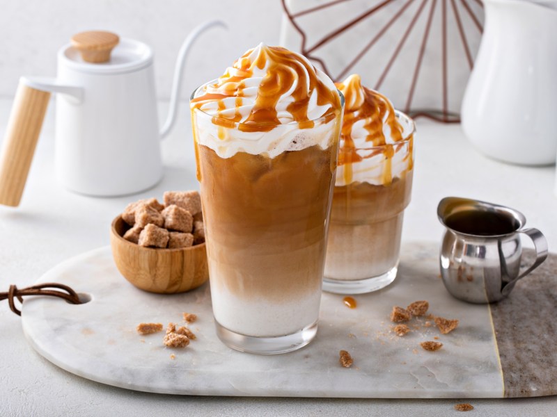 Zwei Gläser Coconut Caramel Latte mit Schlagsahne und Sirup garniert auf einem kleinen Tablett. Im Hintergrund eine Kaffeekanne, daneben ein Espressokännchen und eine Zuckerschale.