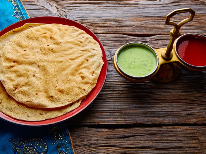 Mehrere Papadams auf einem Teller. Daneben stehlen Schalen mit Soße.