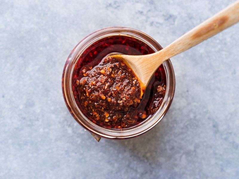 Draufsicht auf ein Glas Salsa Macha mit einem Holzlöffel darin. Das Glas mit der Salsa Macha steht auf einem hellgrauen Untergrund.