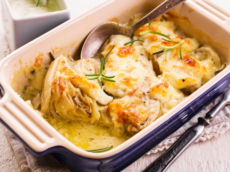 Eine weiß-dunkelblaue rechteckige Auflaufform mit überbackenem Chicorée garniert mit frischem Rosmarin. Die Auflaufform steht auf einem Tuch auf hellem Untergrund. Rechts daneben liegt eine Gabel. In der linken oberen Bildecke kann man verschwommen eine kleine quadratische Schale erkennen.