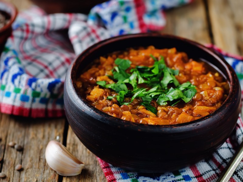 Eine Schüssel veganer Linseneintopf mit Kräutern garniert, drumherum Knoblauch und ein Geschirrtuch.