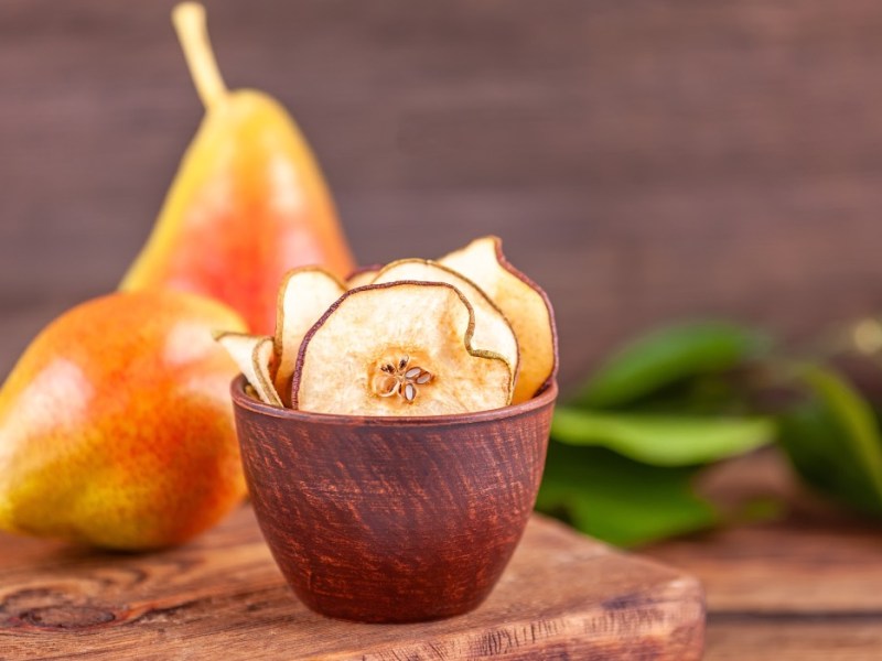 Ein Schälchen mit Birnenchips. Im Hintergrund liegen zwei frische Birnen.