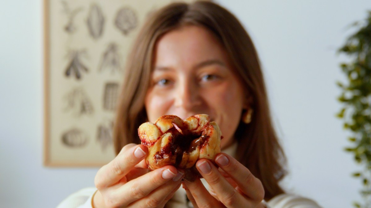 Unsere Köchin hält einen auseinandergezogenen Brioche-Muffin in die Kamera. Man sieht deutlich die Schokoladenfüllung. Im Hintergrund hängt ein Bild mit verschiedenen Wurzelgewächsen.