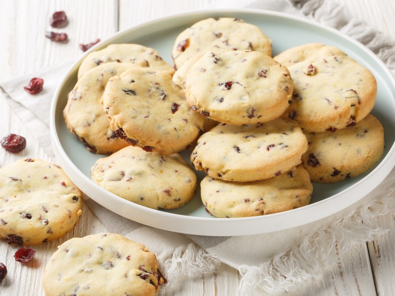 Ein weißer Teller mit einigen Cranberry-Orange-Shortbread-Cookies. Der Teller steht auf einem kleinen weiß-grau gestreiften Küchentuch. Neben den Teller liegen zwei einzelne Kekse und einige Cranberrys verstreut.