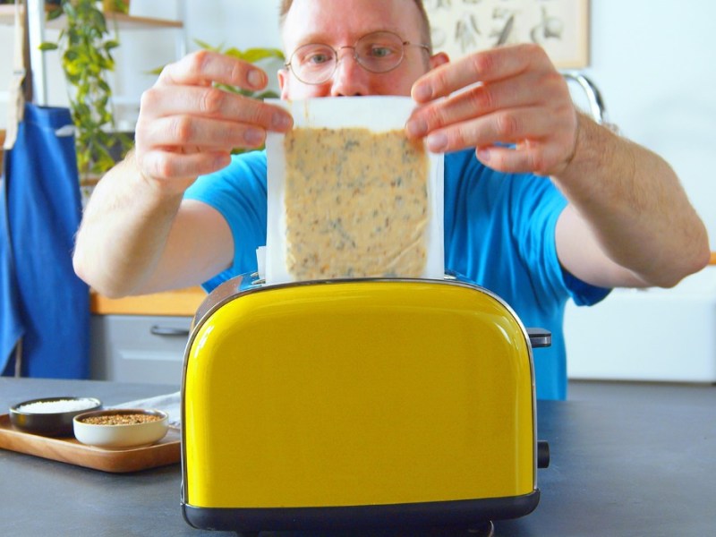 Fetabrotteig für ein Feta-Sandwich wird zwischen zwei Backpapieren in einen Toaster gegeben.