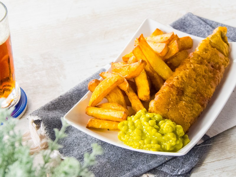 Fish and Chips in einer Schale, auf Zeitungspapier angerichtet. Dabei Erbsenpüree und Zitrone.