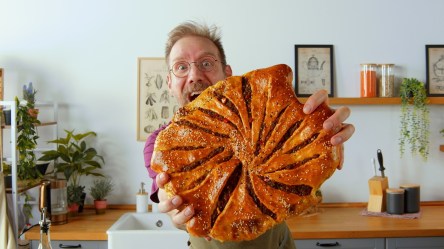 Koch Oli präsentiert ein großes selbst gemachtes und knusprig goldbraun gebackenes Hack-Fladenbrot. Im Hintergrund ist eine Küche zu sehen.