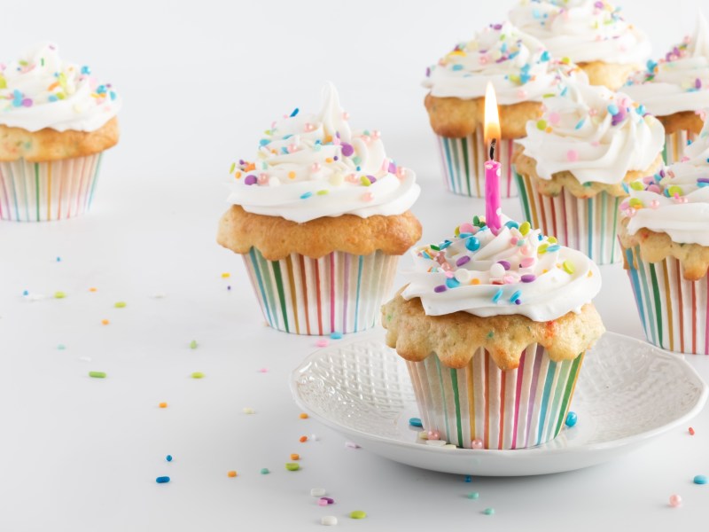 Mehrere Konfetti-Cupcakes mit Zuckerstreuseln verziert, in einem steckt eine brennende Kerze.