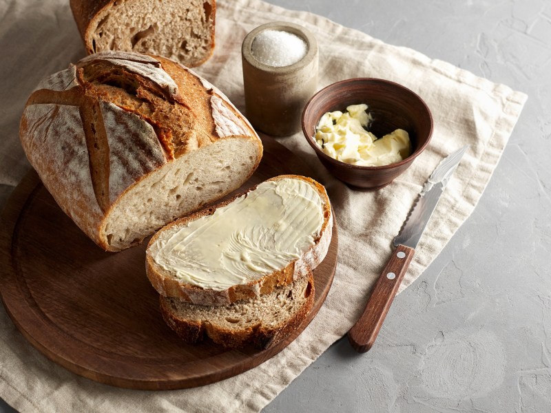 Landbrot auf einem Holzbrett. Dieses steht auf einer sauberen Serviette. Von dem Brot wurden mehrere Scheiben abgeschnitten, eine wurde mit Butter bestrichen.