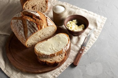 Landbrot auf einem Holzbrett. Dieses steht auf einer sauberen Serviette. Von dem Brot wurden mehrere Scheiben abgeschnitten, eine wurde mit Butter bestrichen.