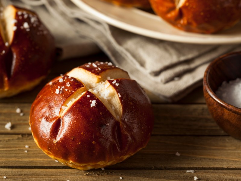 Ein Laugenbrötchen vor einem Teller mit weiteren Laugenbrötchen, den man nur angeschnitten sieht. Nahaufnahme.