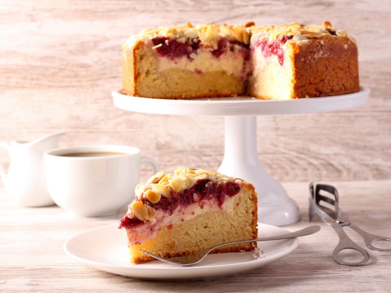 Mandelkuchen mit Quark und Kirschen auf einem Kuchentablett. Ein Stück wurde herausgeschnitten.
