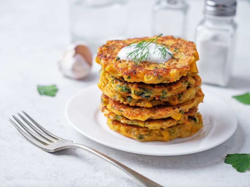Ein Stapel Pastinaken-Möhren-Puffer mit Dill und Petersilie. Obendrauf ist ein Klecks Joghurts.