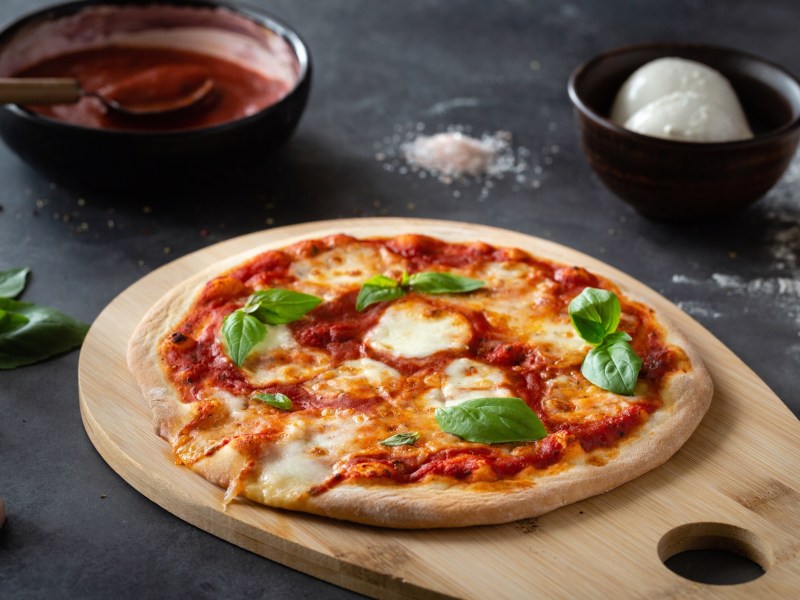 Eine Pizza Margherita mit Tomatensoße, Mozzarella und Basilikum auf einem großen Holzbrett, drumherum einige Zutaten