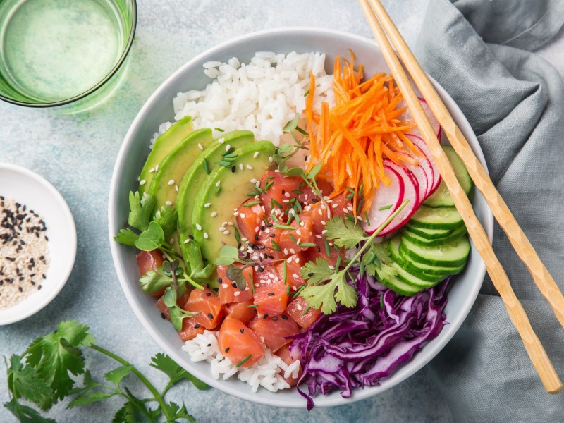 Poke Bowl mit frischem Fisch, Gemüse und Sesam in der Draufsicht. Daneben liegen einige der Zutaten.