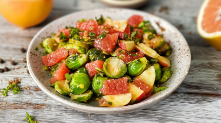 Fruchtiger Rosenkohlsalat in einer hellgrauen Schüssel. Er besteht aus Rosenkohl, Grapefruitstückchen und Apfelstücken. Die Schüssel steht auf einem Holztisch. Im Hintergrund liegen noch einige Grapefruits.